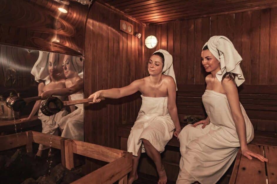 Two women in towels pouring water in a wood-fired steam sauna